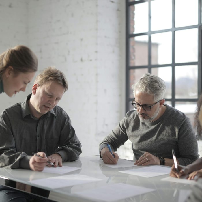 Female supervisor watching diverse workers of different ages writing on papers while gathering at table in light studio